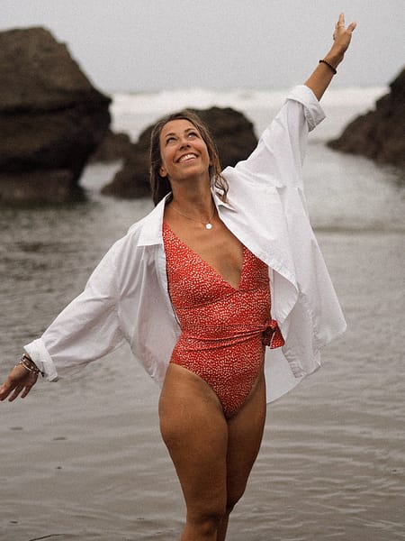 Une femme bras écartés sur une plage. Elle est en maillot de bain une pièce rouge à point blanc avec par dessus une chemise blanche ouverte.